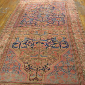 Ornate low-pile rug in rose, navy blue, and sage green displayed on light wood floorboards.