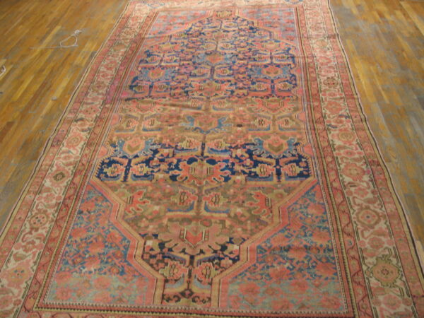 Ornate low-pile rug in rose, navy blue, and sage green displayed on light wood floorboards.