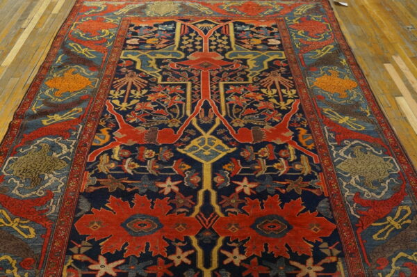 Ornate traditional rug featuring a dark blue field, red central elements, and stylized figures on wood flooring.