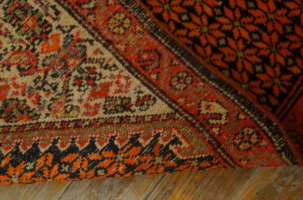 A geometric rug in red, orange, black, and tan is partially rolled back on distressed wood flooring.