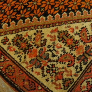 Ornate rug corner with orange geometric field, tan border, red motifs, and visible fringe on wood planks.