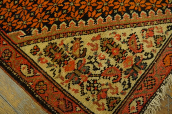 Ornate rug corner with orange geometric field, tan border, red motifs, and visible fringe on wood planks.