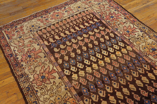 A low-pile rug with a dark brown field, blue/rose motifs, and a wide floral beige border, lying on wood floors.