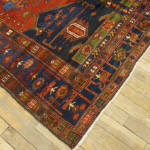 A corner of a red, navy, and green geometric patterned rug rests on rustic light wood floorboards.