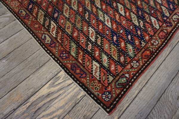 Red and navy geometric diamond pattern rug corner rests on distressed gray hardwood flooring.