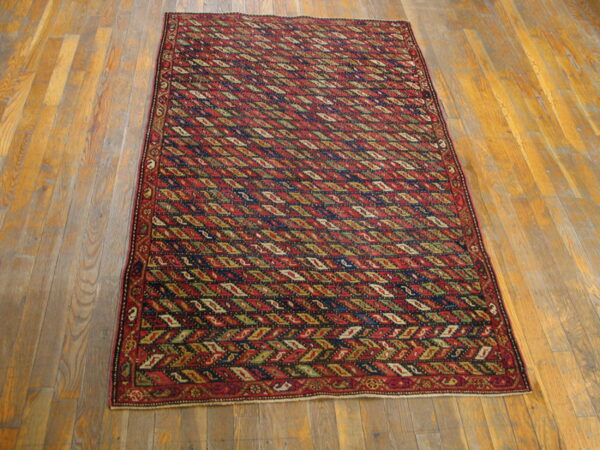 Red and navy geometric rug with repeating olive and cream diamonds resting on wood plank flooring.