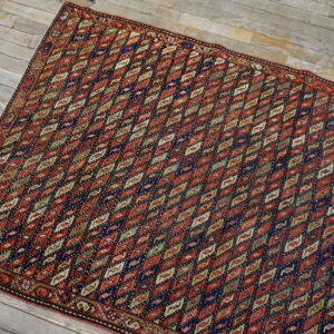 Red and navy low pile rug with repeating multicolored diamonds rests on distressed gray wood floors.