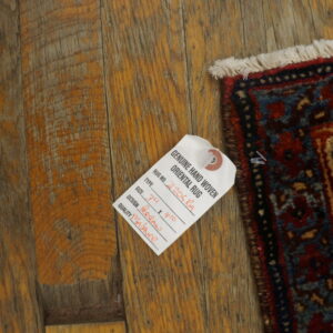 Corner of a red and blue patterned rug with fringe on distressed wood flooring with an attached tag.