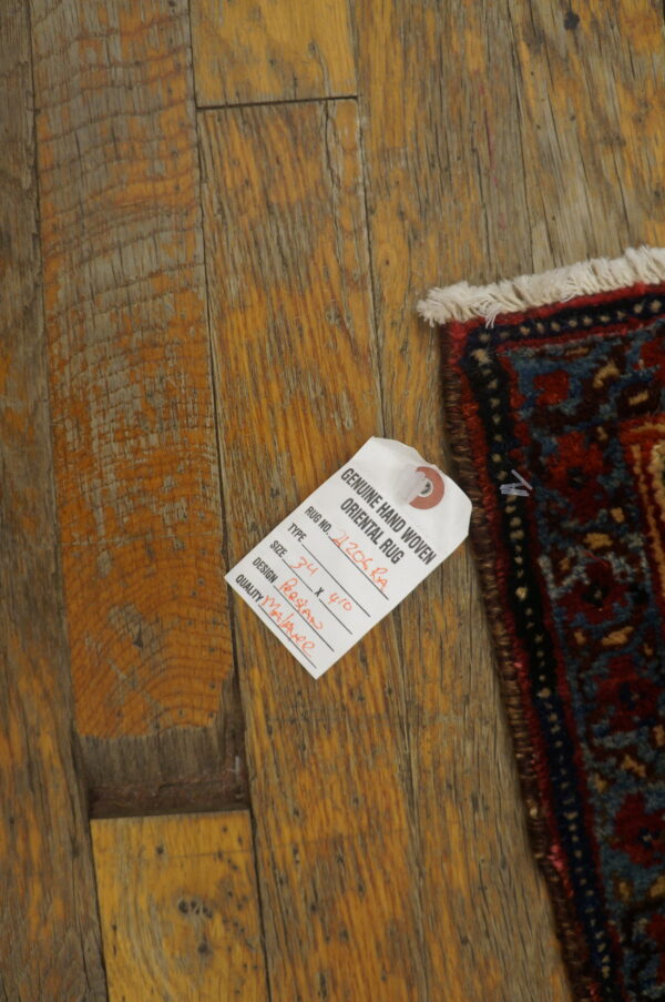 Corner of a red and blue patterned rug with fringe on distressed wood flooring with an attached tag.