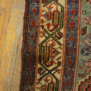 Close-up of a traditionally patterned rug with red, blue, green, and beige accents beside light wood flooring.