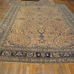 Tan area rug with complex blue floral medallion and borders rests on light wood plank flooring.