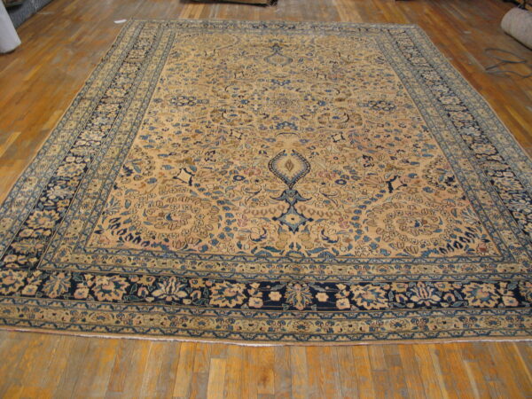 Tan area rug with complex blue floral medallion and borders rests on light wood plank flooring.