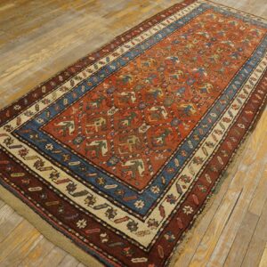A rust red rug with tribal geometric patterns and a blue and cream border lies on light wood planks.