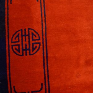 Red rug detail featuring a navy blue medallion design and border, set against light brown wooden flooring.