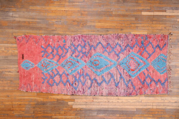 Vibrant pink high-pile rug featuring blue geometric diamonds and zigzags displayed on hardwood flooring.