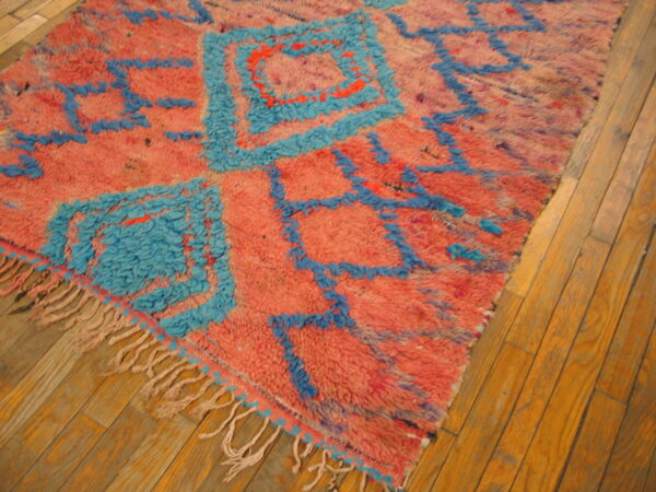High-pile pink rug with abstract bright blue geometric designs and fringes rests on a wooden floor.