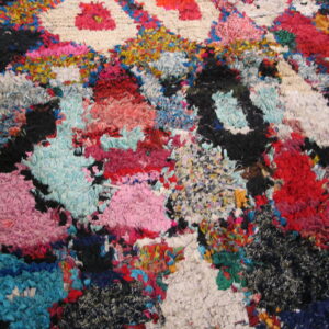 A close-up shows a highly textured, abstract rug with bright pink, blue, red, and black fabric scraps on wood flooring.