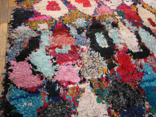 A close-up shows a highly textured, abstract rug with bright pink, blue, red, and black fabric scraps on wood flooring.