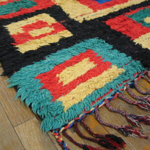 Shaggy rug featuring geometric blocks of red, yellow, black, and teal, with braided fringe on wood.
