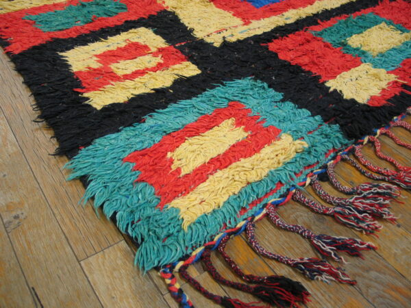 Shaggy rug featuring geometric blocks of red, yellow, black, and teal, with braided fringe on wood.