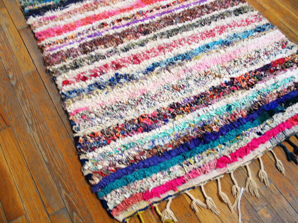 Chunky, striped multi-color rag rug with fringe detail resting on warm brown wood flooring.