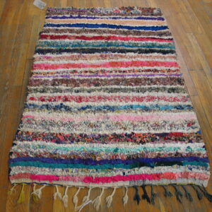 Textured, multicolored rag rug with horizontal stripes and fringe rests on a warm wood floor.