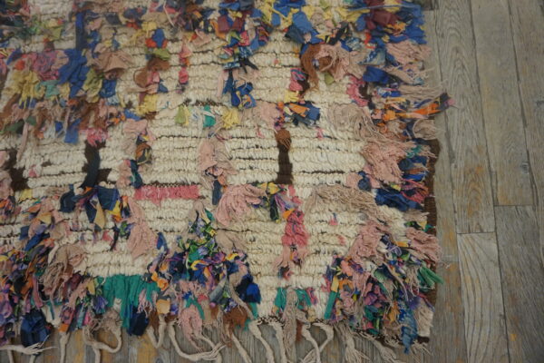 High-pile, textured cream rug with colorful knotted fabric strips lying on weathered gray wood floor.