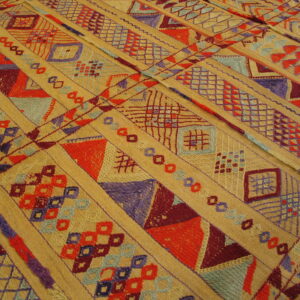 Golden rug with dense diagonal stripes of embroidered red, purple, and blue geometric motifs on low pile.