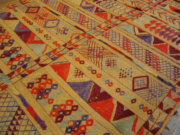 Golden rug with dense diagonal stripes of embroidered red, purple, and blue geometric motifs on low pile.