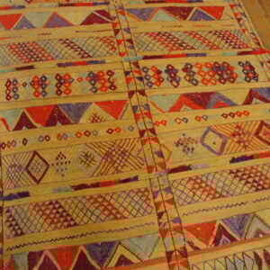 Flat-woven rug displaying horizontal bands of geometric motifs in tan, red, purple, and light blue on wood flooring.