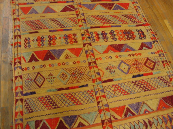 Flat-woven rug displaying horizontal bands of geometric motifs in tan, red, purple, and light blue on wood flooring.