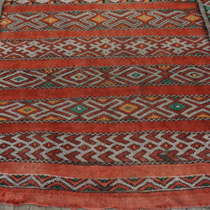 Coarse, red flat-weave rug featuring geometric stripes in white, green, and yellow on rustic wood flooring.