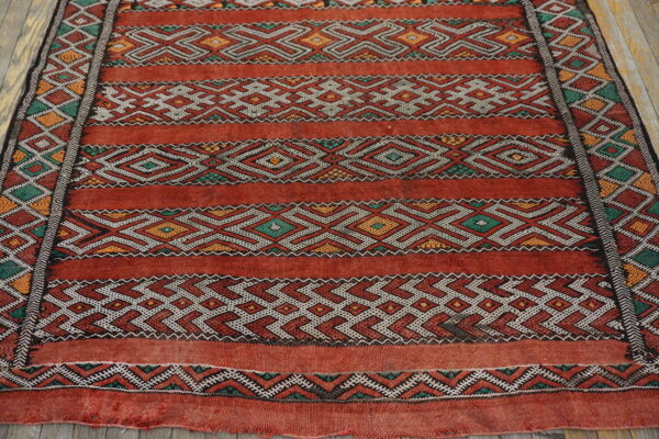 Coarse, red flat-weave rug featuring geometric stripes in white, green, and yellow on rustic wood flooring.