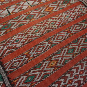 Red low-pile rug with diagonal bands of white, green, and orange geometric diamond and zig-zag patterns.