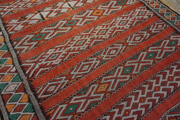 Red low-pile rug with diagonal bands of white, green, and orange geometric diamond and zig-zag patterns.
