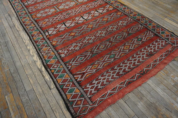 Rust-toned flatweave rug featuring geometric bands of white, emerald, and orange on weathered wood planks.