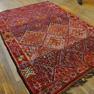 Shaggy red rug featuring multicolored diamonds and geometric motifs covers a light brown wood floor.