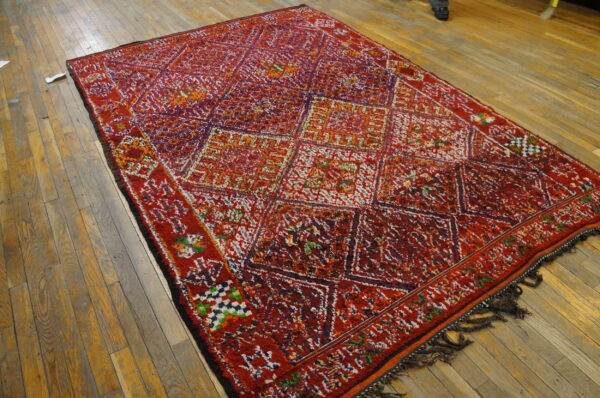 Shaggy red rug featuring multicolored diamonds and geometric motifs covers a light brown wood floor.