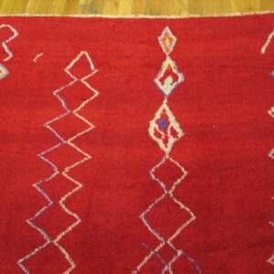 Textured red rug with white and blue geometric diamonds displayed on wood flooring.