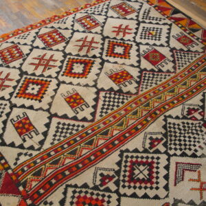Cream high-pile rug with geometric patterns in black, red, orange, and yellow on distressed wood floor.