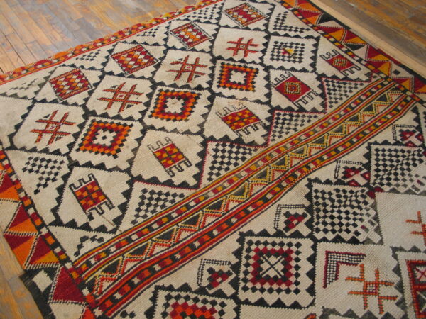 Cream high-pile rug with geometric patterns in black, red, orange, and yellow on distressed wood floor.