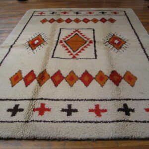 Shaggy off-white rug with geometric orange, red, and dark brown diamond and cross patterns on wood flooring.