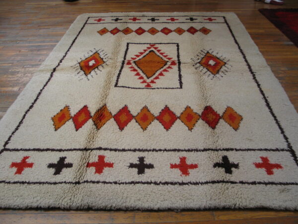 Shaggy off-white rug with geometric orange, red, and dark brown diamond and cross patterns on wood flooring.