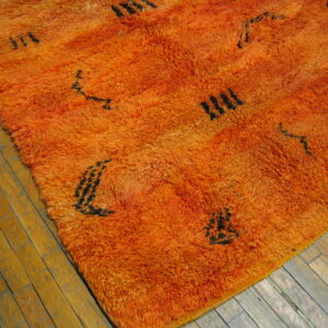 High-pile orange rug featuring sparse black abstract symbols rests on worn light wood plank flooring.