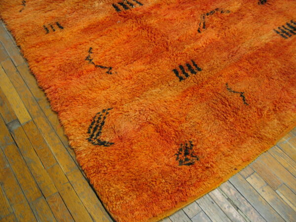 High-pile orange rug featuring sparse black abstract symbols rests on worn light wood plank flooring.