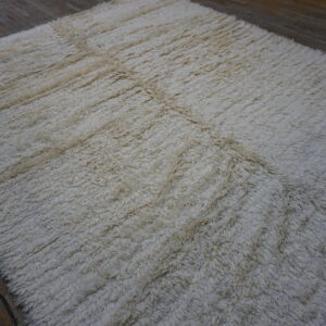 A high-pile, off-white rug featuring a faint geometric grid rests on dark, distressed wood plank flooring.