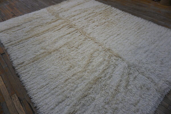 A high-pile, off-white rug featuring a faint geometric grid rests on dark, distressed wood plank flooring.