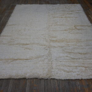 Shaggy, off-white rectangular rug with high pile covering dark brown plank flooring.