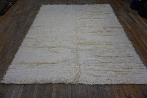 Shaggy, off-white rectangular rug with high pile covering dark brown plank flooring.