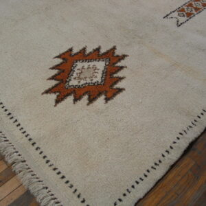 Textured cream rug with orange and black geometric pattern and fringe over dark wood floor.
